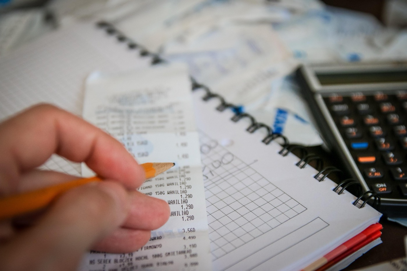 Hand mit Bleistift, Kassenzettel, kariertes Notizheft und Taschenrechner auf einem Tisch